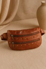 Cognac brown leather waist pouch with brass studs and dual zippers by Dario Lario on neutral backdrop