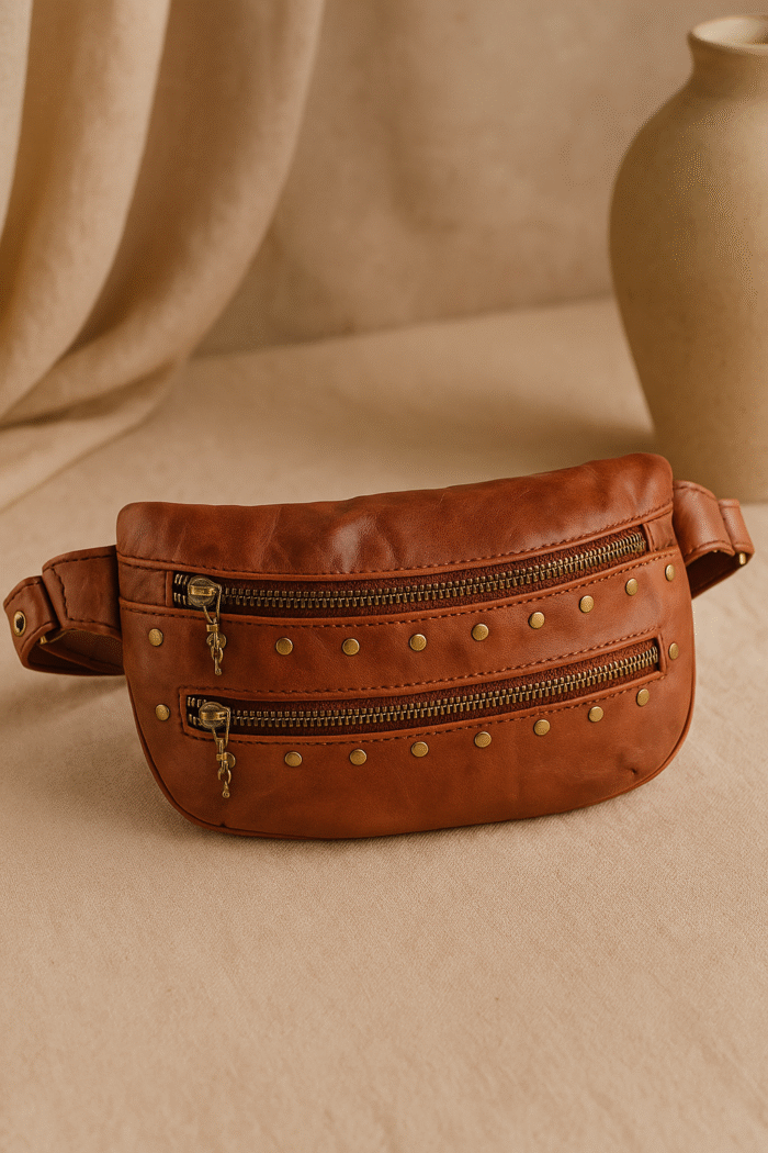 Cognac brown leather waist pouch with brass studs and dual zippers by Dario Lario on neutral backdrop